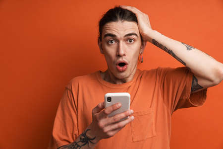 Young white man expressing surprise while using cellphone isolated over orange background