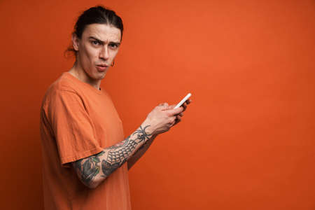 Young white man expressing surprise while using cellphone isolated over orange background