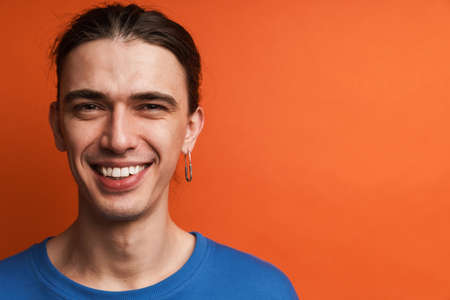 Young white man with earring looking and laughing at camera isolated over orange background