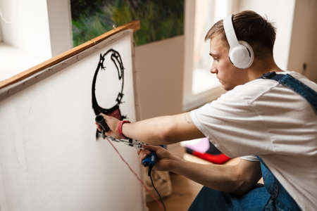 Young white man in headphones working on craft rug with sewing machine at homeの写真素材