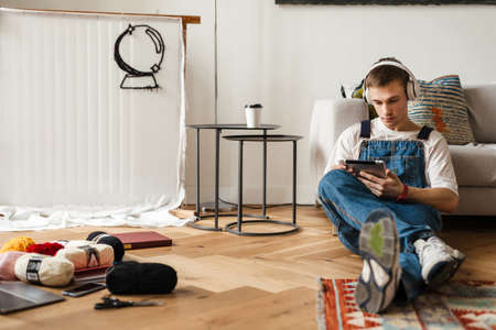 Young man in headphones using tablet computer while working on craft rug at home