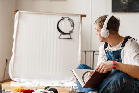 Young man in headphones writing down notes while working on craft rug at home