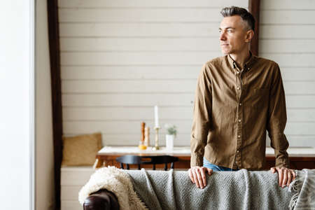 Smiling middle aged white man standing in the modern living room, leaning on a couch, looking asideの写真素材