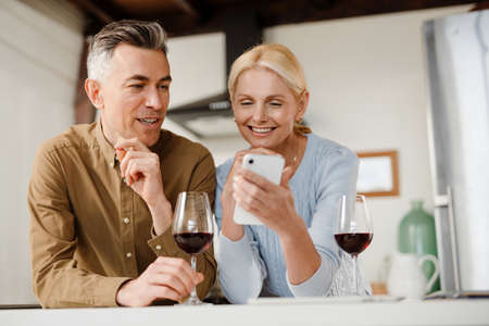 Smiling middle aged couple standing together at the kitchen drinking red wine at home looking at mobile phoneの写真素材