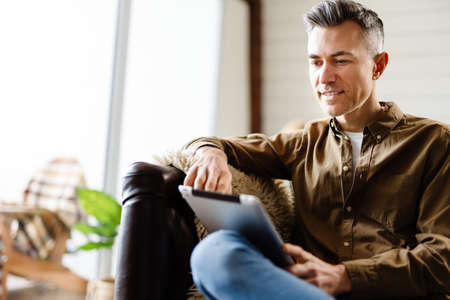 Smiling mid aged white man holding tablet computer sitting on a sofa indoorsの写真素材