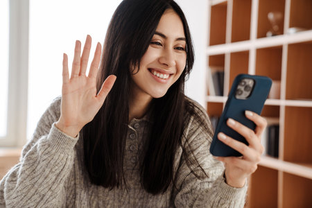 Happy young woman on a video call via mobile phone while at home, waving handの写真素材