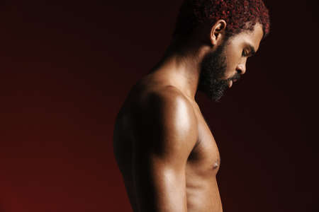 Black shirtless bearded man posing and looking downward isolated over red backgroundの写真素材