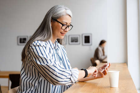 Smiling white-haired mature woman looking at smartwatch and using cellphone indoorsの写真素材