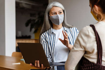 Two employee standing in social distance wearing face mask talking in the officeの写真素材