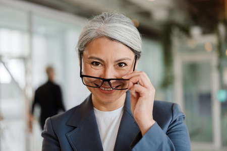 Happy white-haired mature businesswoman in eyeglasses smiling at camera in officeの写真素材