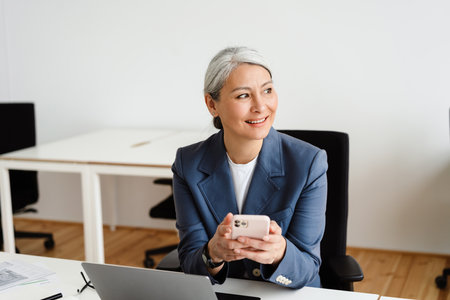 Happy white-haired businesswoman smiling while working with laptop and cellphone in officeの写真素材