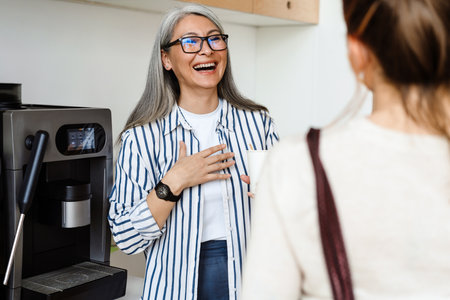 Happy white-haired mature woman drinking coffee while talking with her colleague indoorsの写真素材