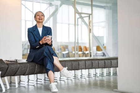 Cheerful white-haired businesswoman using cellphone and laughing in officeの写真素材