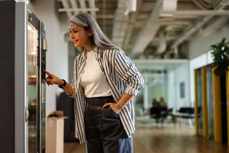 Happy white-haired mature woman using vending machine and smiling indoorsの写真素材
