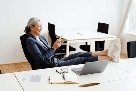 Laughing white-haired businesswoman using cellphone while working with laptop in officeの写真素材