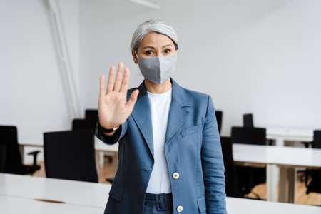 Confident white-haired mature businesswoman in face mask showing stop gesture at officeの写真素材