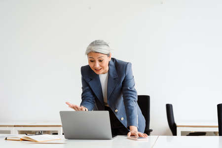Emotional white-haired businesswoman gesturing while conference call on laptop in officeの写真素材