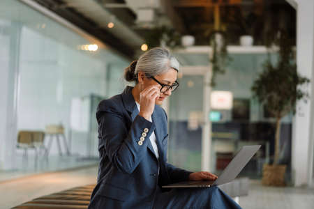 Focused white-haired businesswoman using laptop while sitting in officeの写真素材