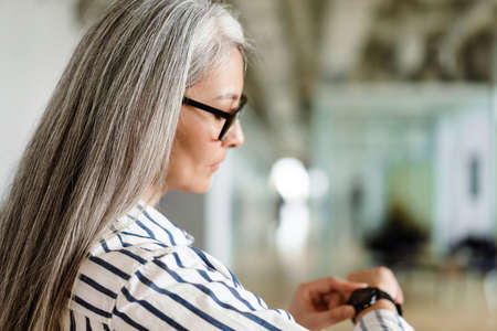 Focused white-haired mature woman in eyeglasses using her wristwatch indoorsの写真素材