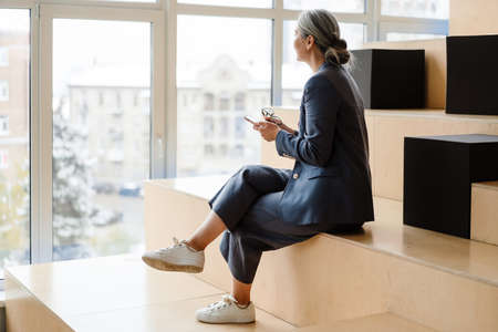 Elegant white-haired businesswoman using cellphone while sitting indoorsの写真素材