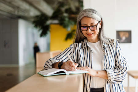 Joyful white-haired mature woman smiling while writing down notes indoorsの写真素材