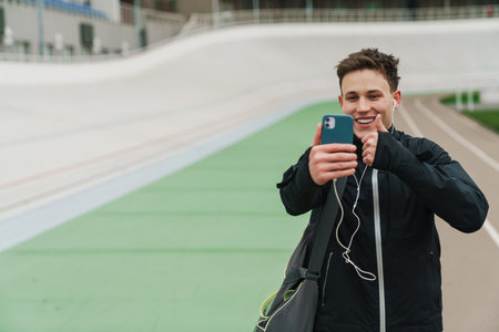 Happy sportsman in earphones showing thumb up while taking selfie on cellphone at stadium outdoorsの写真素材