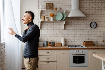 Handsome pleased man smiling and looking out window at home kitchenの写真素材