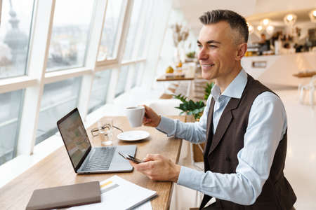 Confident handsome businessman in smart clothes working on laptop computer while sitting in the cafe indoors, using mobile phone, drinking coffeeの写真素材