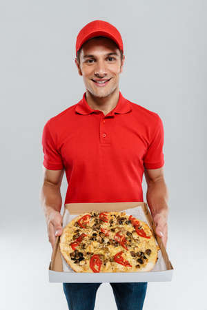 Young delivery man in uniform smiling while showing pizza isolated over white wallの写真素材