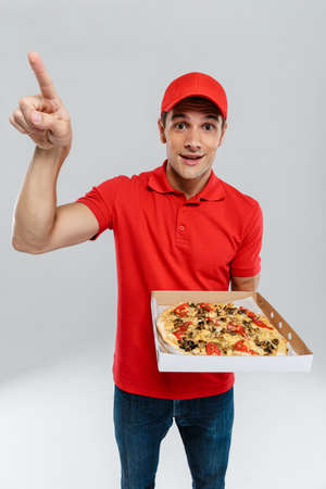 Young delivery man pointing finger upward while showing pizza isolated over white wallの写真素材