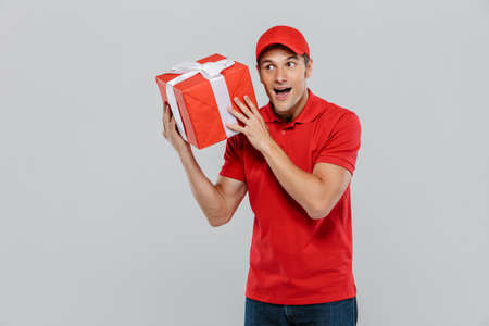 Young delivery man in hat smiling while posing with gift box isolated over white wallの写真素材