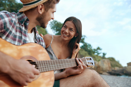 Happy guy play on guitar to beautiful smiling girl. Young european couple in love resting on sandy beach. Concept of romantic relationship and enjoying time together. Vacation on nature at summertimeの写真素材