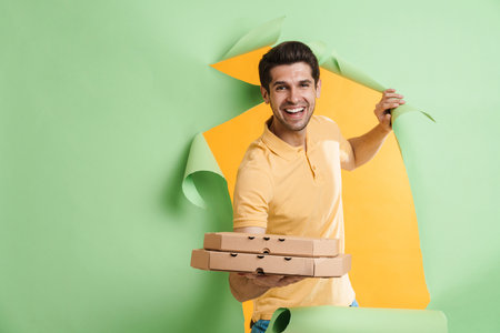 Young unshaven man smiling while posing with pizza boxes into hole in green backgroundの写真素材