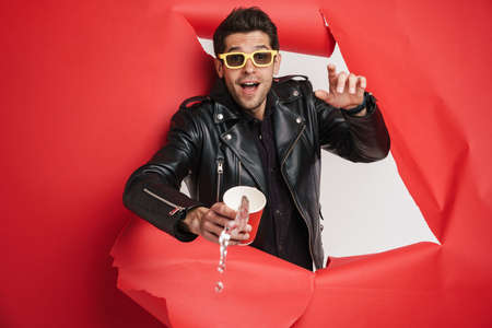 Young unshaven man in sunglasses pointing finger at camera while posing into hole in red background