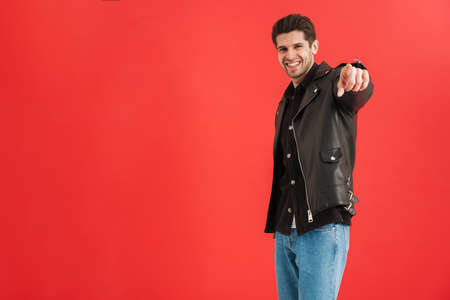 Young man wearing leather jacket pointing finger at camera isolated over red wall