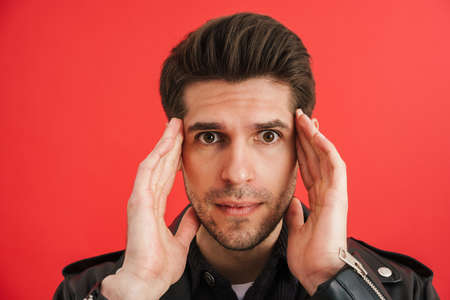 Young unshaven man in leather jacket expressing surprise at camera isolated over red wallの写真素材