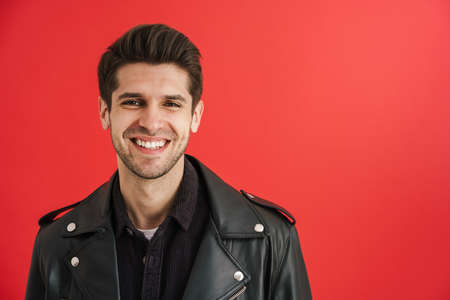 Young unshaven man wearing leather jacket smiling at camera isolated over red wallの写真素材