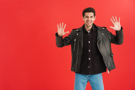 Young man wearing leather jacket smiling and gesturing at camera isolated over red wall