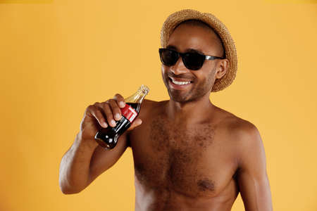Young smiling handsome black man drinking soda from bottle. Guy wear swimming trunks, straw hat and glasses. Concept of summer beach vacation. Isolated on yellow background. Studio shootの写真素材