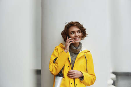 Young girl in yellow raincoat calling on smartphone while walking in the city. Young beautiful brown...