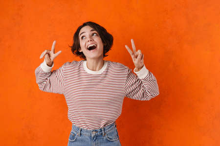 Young brunette woman screaming while showing peace gesture isolated over orange wall