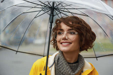 Young girl in yellow raincoat with transparent umbrella in city. Young beautiful woman wearing rain...