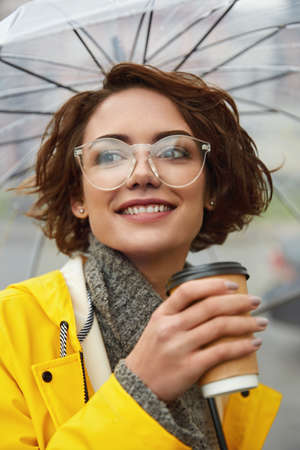 Young girl in yellow raincoat with transparent umbrella in city. Young beautiful woman wearing rain...