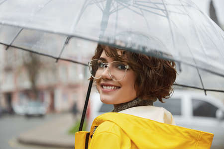 Young girl in yellow raincoat with transparent umbrella in city. Young beautiful woman wearing rain outfit and eyeglasses while walking on street. Concept of modern woman lifestyle at autumn.の写真素材