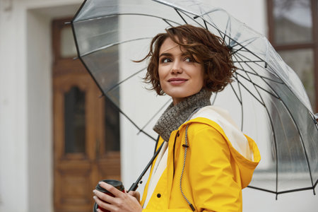 Caucasian girl in yellow raincoat with transparent umbrella in city. Young beautiful woman wearing rain outfit while walking on street at daytime. Concept of modern woman lifestyle at autumn season.の写真素材