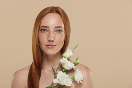 Ð¡aucasian girl with naked shoulders holds eustoma flower. Cropped partial view of young beautiful red haired woman with freckles. Concept of natural female beauty. Isolated on beige backgroundの写真素材