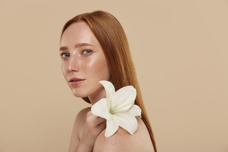 Redhead caucasian girl with naked shoulders holds lily flower. Cropped view of young beautiful red haired woman with freckles. Concept of natural female beauty. Isolated on beige background in studioの写真素材