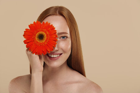 Redhead caucasian girl with naked shoulders holds gerbera flower. Cropped partial view of young beautiful red haired woman with freckles. Concept of natural female beauty. Isolated on beige backgroundの写真素材