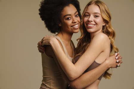 Two diverse young women in underwear standing over beige background close up