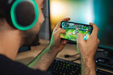 Young man gamer playing video games on smartphone and computer in dark room wearing headphonesの写真素材